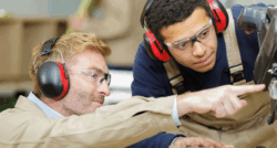 Two people wearing protective earmuffs and work overalls are operating machinery together. One person is pointing toward a part of the equipment while the other observes closely.